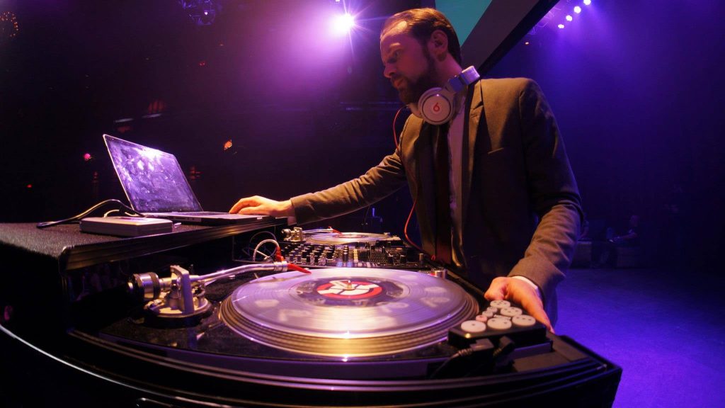 DJ in a suit performing on turntables and laptop under purple stage lighting at a NYX party