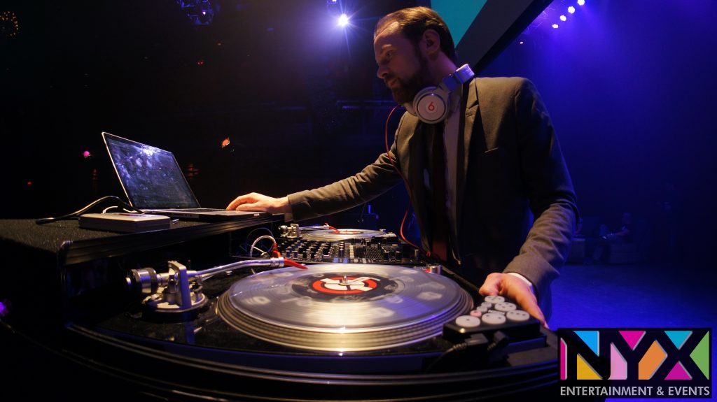 DJ in a suit mixing music on turntables and laptop under stage lights at an NYX Entertainment & Events party