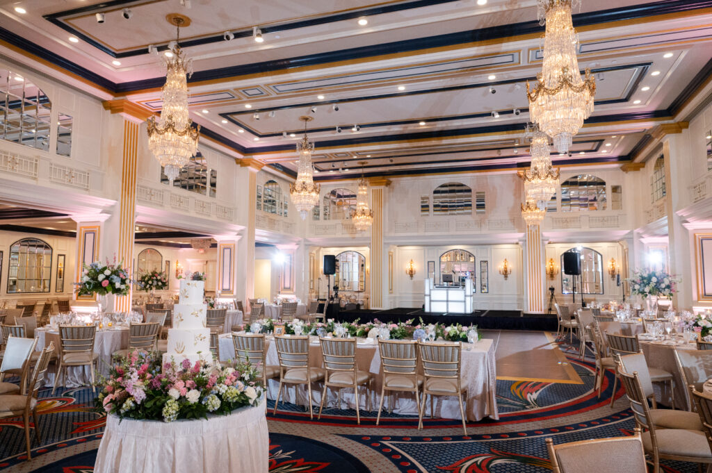 Elegant wedding reception ballroom with chandeliers, floral centerpieces, and a tiered cake surrounded by decorated tables