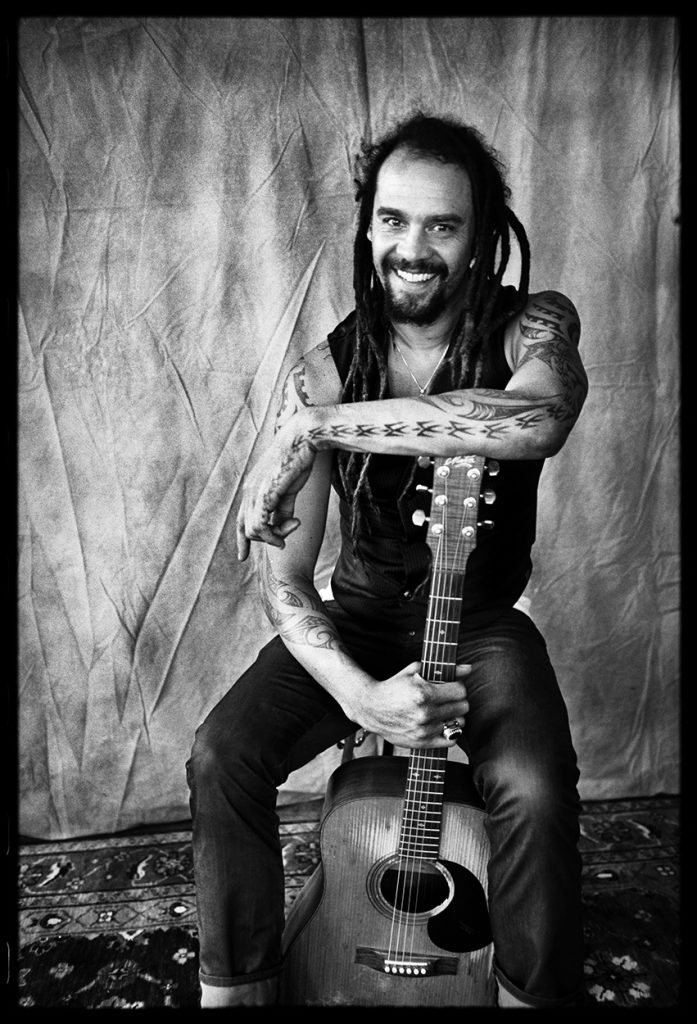 Black and white portrait of musician Michael Franti smiling while seated with an acoustic guitar.