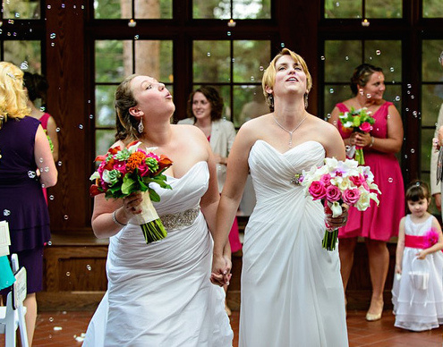 Two brides holding hands and smiling while walking down the aisle with bouquets during their wedding ceremony