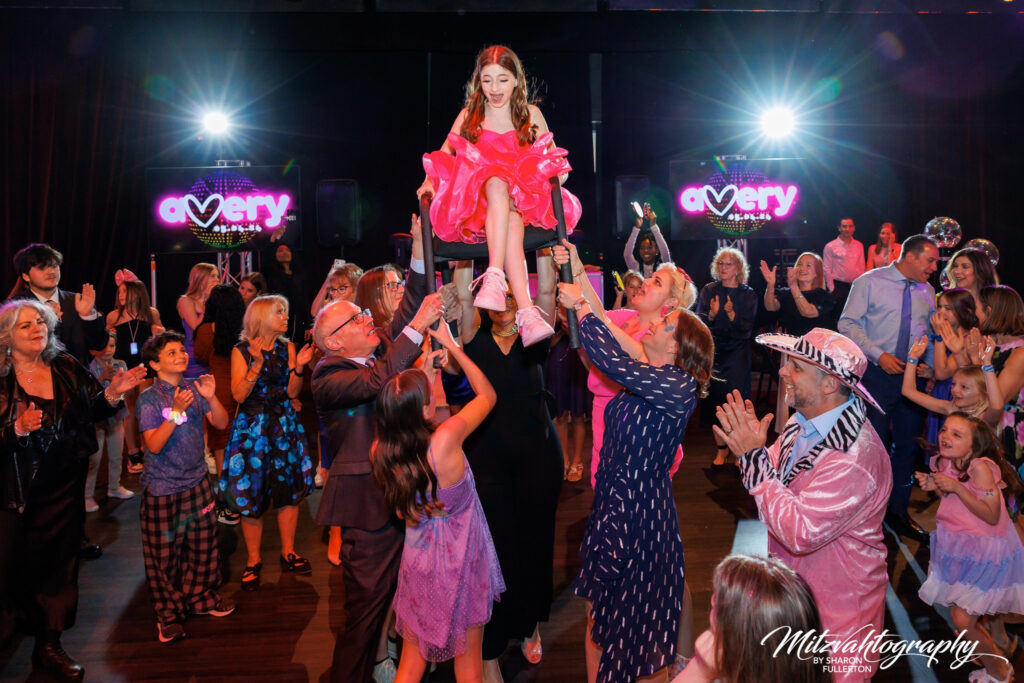 Young girl lifted in a chair during the Hora dance at her Bat Mitzvah celebration, surrounded by cheering guests.