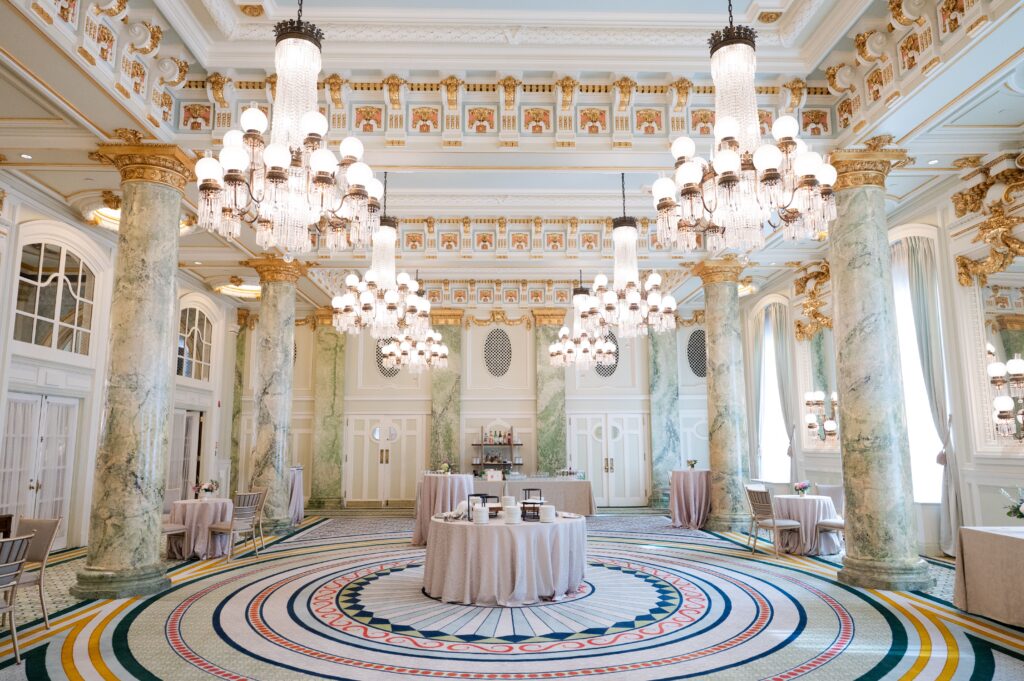 Elegant ballroom with marble columns, ornate gold detailing, large crystal chandeliers, and round tables draped in light linens, set for a wedding reception.