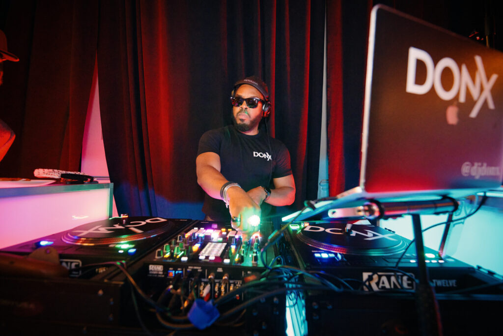 A DJ in sunglasses and headphones mixing live on turntables and a mixer at an event, creating high-energy music for the crowd.