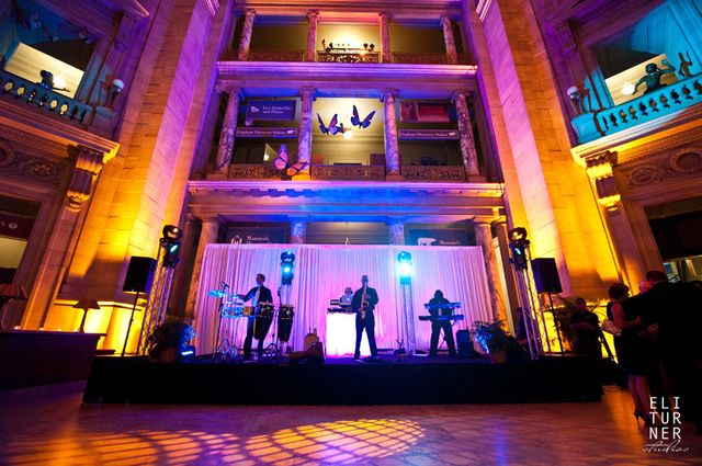 NYX Live wedding entertainment setup with DJ, live horns, and percussionists performing on stage in a grand venue with colorful lighting, showcasing the fusion of live music and DJ energy.