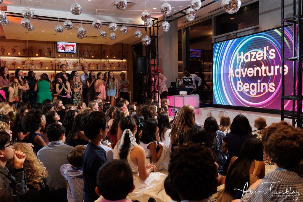 Guests gathered around a large LED screen watching a mitzvah montage that reads “Hazel’s Adventure Begins,” during a celebration produced by NYX Events, highlighting the 7–10 minute sweet spot for perfect montage length.