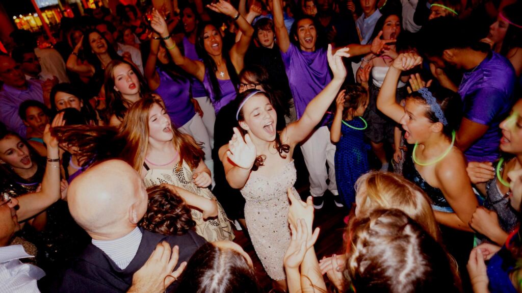 A crowded, high energy dance floor at a Bat Mitzvah celebration with a young girl in a sparkling dress singing and dancing. Guests are wearing purple shirts and neon glow necklaces under vibrant party lighting.