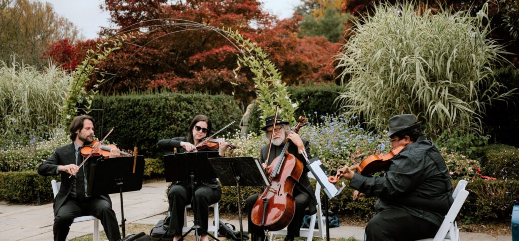Live string act for wedding ceremony playing outdoors in a lush garden under an archway with vibrant red autumn leaves.