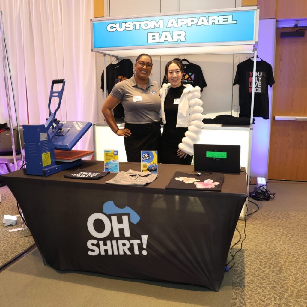 Two women smiling at an interactive mitzvah favors Baltimore booth with a custom apparel bar and heat press.