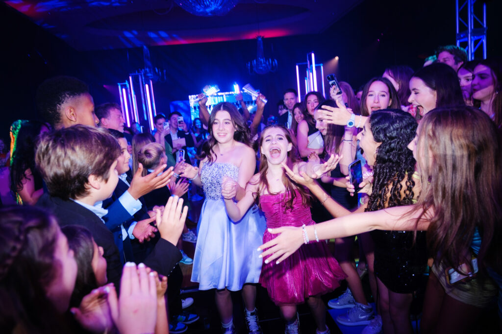A lively crowd of teenagers cheering and reaching out on a dance floor under vivid blue and purple club lighting, focusing on two excited young girls in the center wearing a light blue dress and a shiny pink dress.