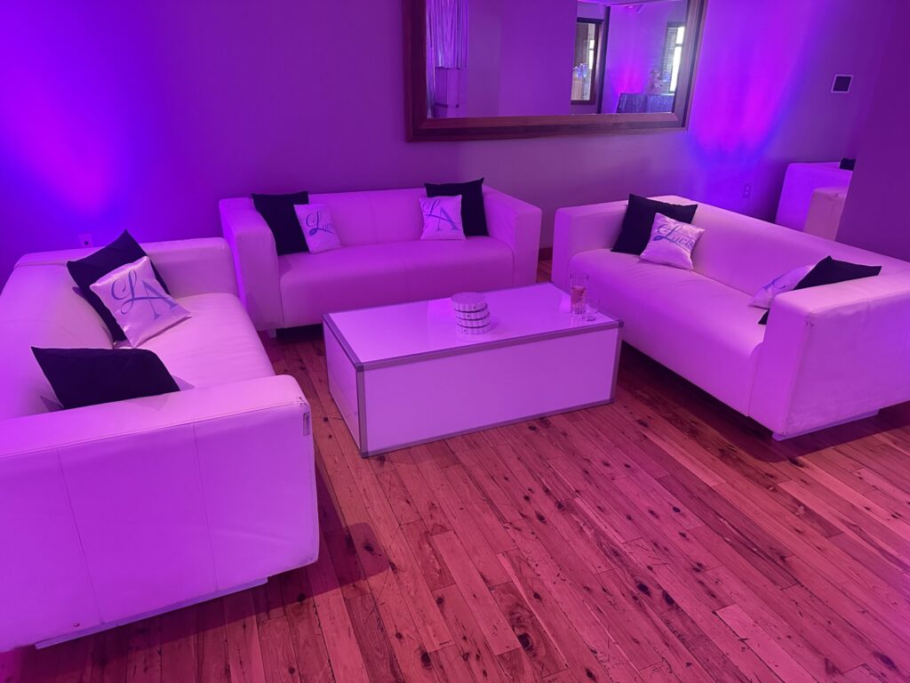 White lounge sofas with personalized pillows arranged around a coffee table under purple uplighting.