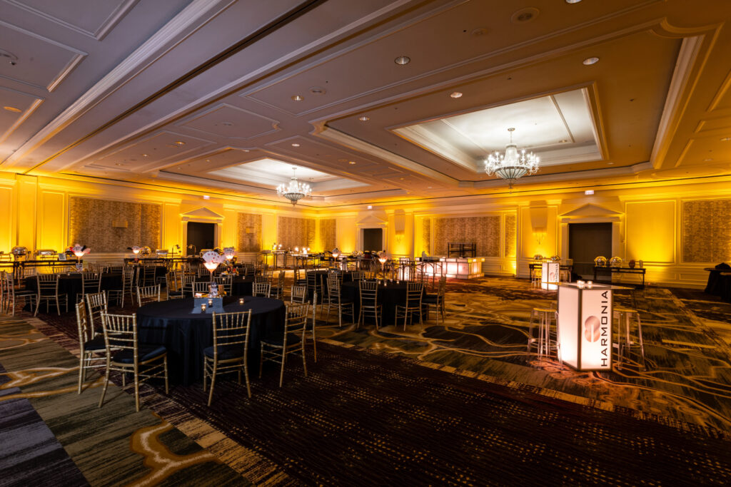 Amber uplighting illuminating ballroom walls, with black-covered tables, gold chairs and glowing cocktail tables.